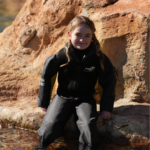 Kira, a child on Dreamflight, sits on a rock with her legs in the water, looking at the camera and smiling. She is wearing a dry suit which helps her to swim without affecting her skin condition.