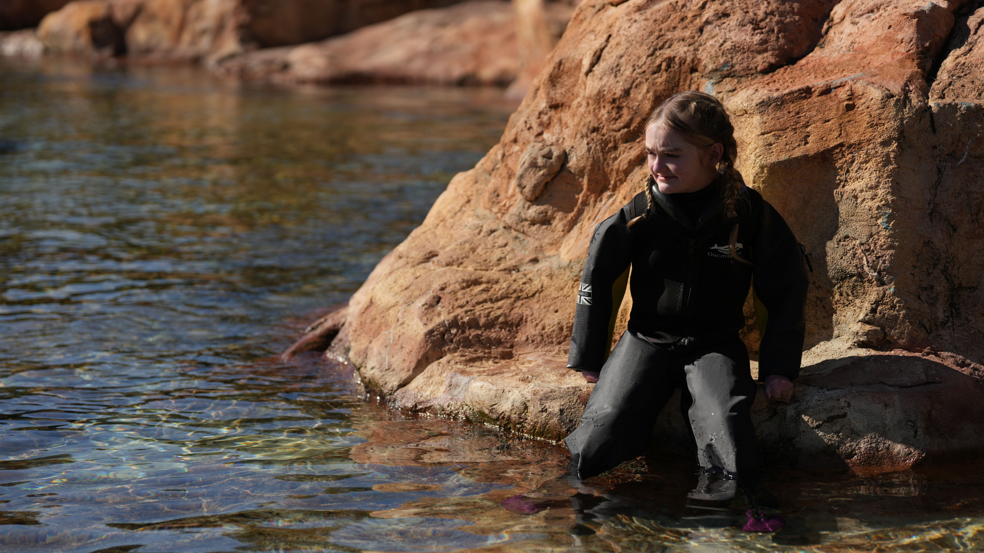 Kira, a child on Dreamflight, sits on a rock looking into the distance with her legs in the water. She is wearing a dry suit which helps her to swim without affecting her skin condition.