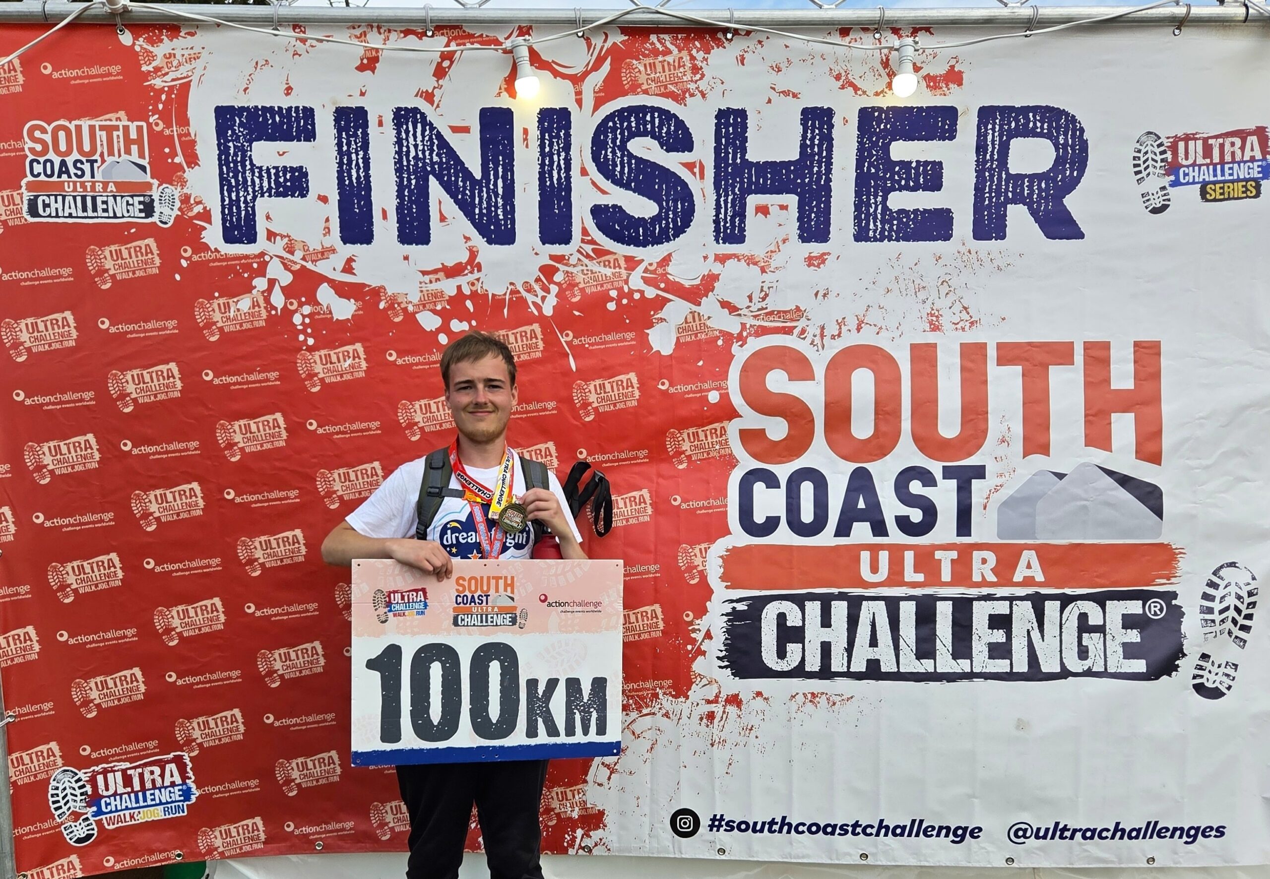 Ryan stood in front of a banner with a medal around his neck and a 100km sign in his hands at the finish line of the South Coast Ultra Challenge.