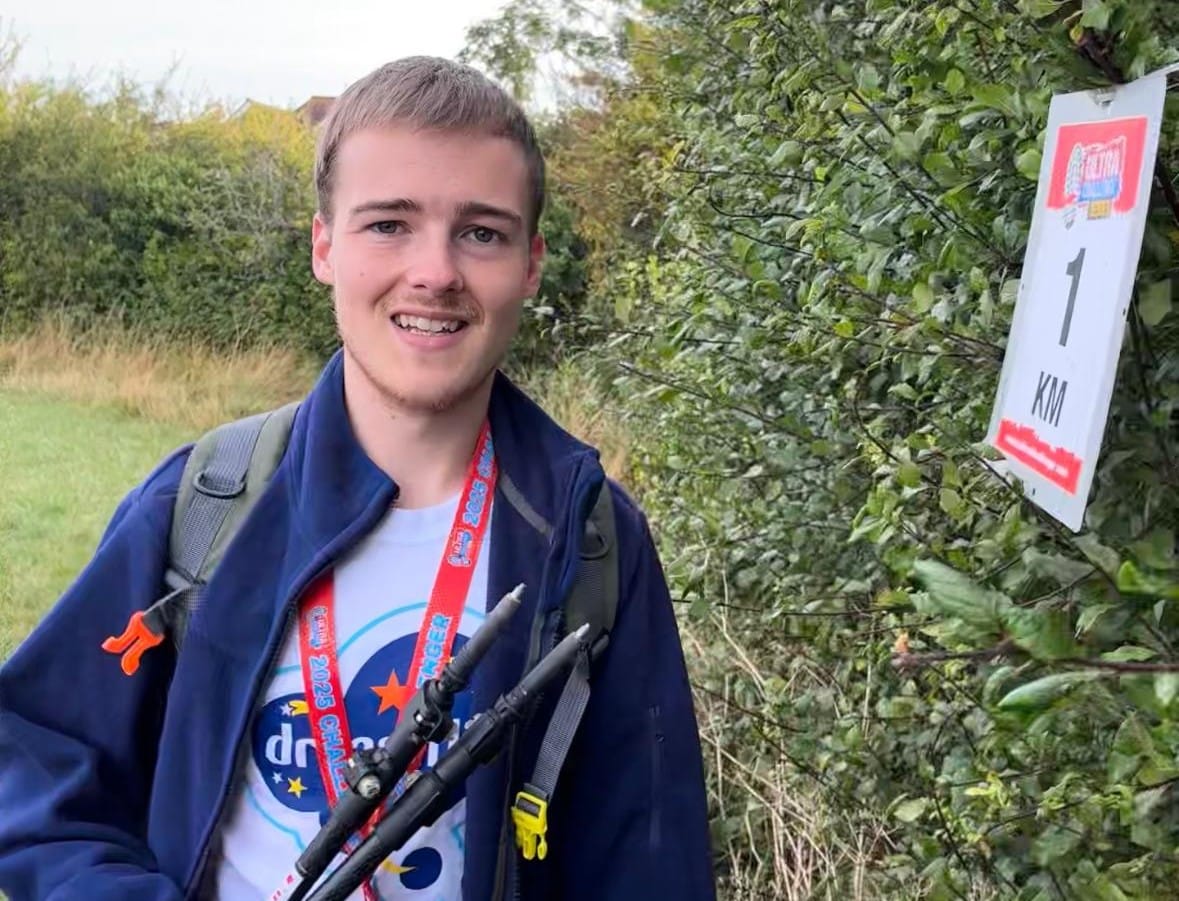 Ryan, smiling for a photo at the 1km mark sign by some bushes, on his ultra challenge.